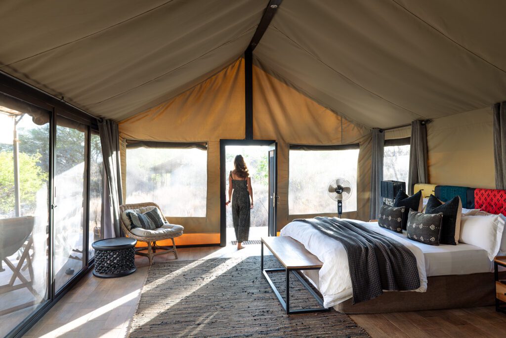 A woman stands at the entrance of a luxury Tanzania lodge tented room with a neatly made bed, armchair, glass walls, and stunning views of nature at Nimali Tarangire.