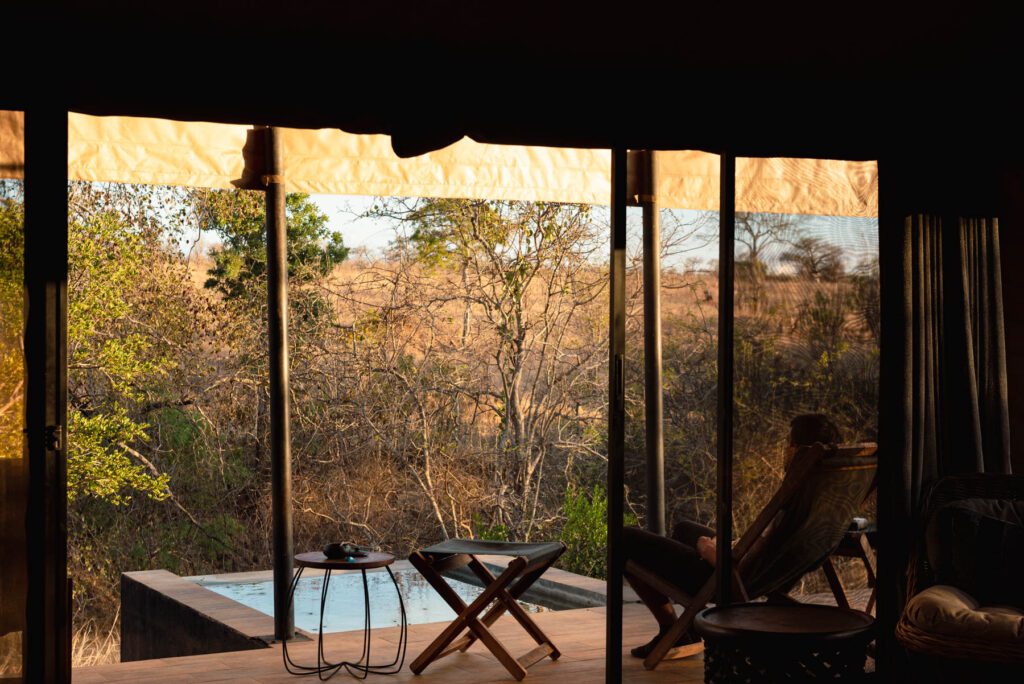 A person sits on a deck chair beside a small pool at Nimali Tarangire, looking out at dry trees and bushes through large glass doors under a shaded canopy.