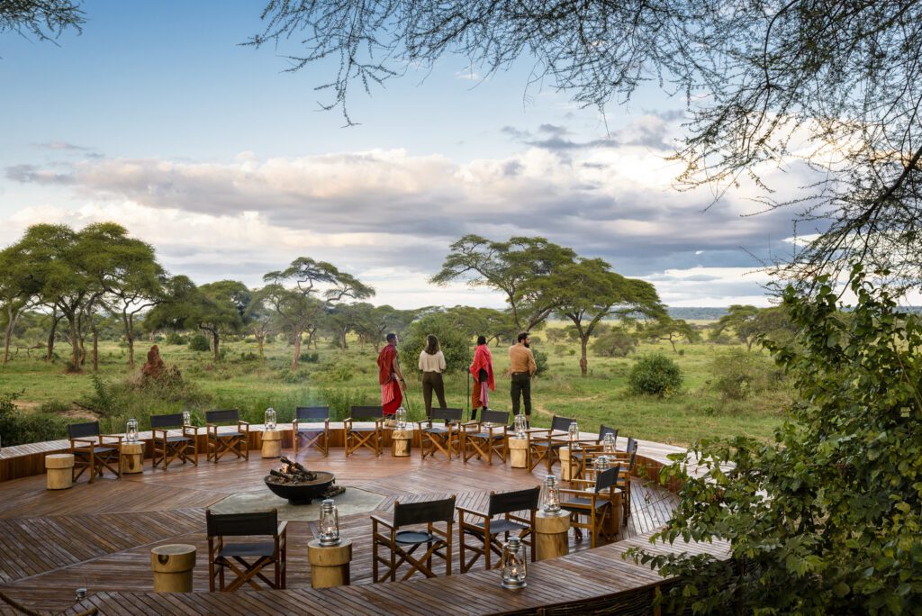 A circular wooden outdoor deck with chairs and lanterns at Sanctuary Swala safari lodge overlooks the green savannah of Tarangire National Park. Four people stand at the edge, facing scattered trees under a cloudy sky.