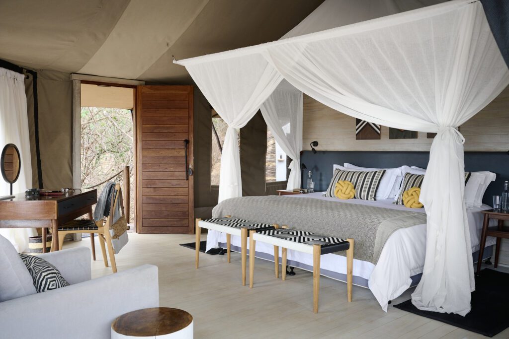 Luxury tented bedroom with a canopy bed, wooden furniture, and modern decor at Sanctuary Swala, featuring an open door with views of Tarangire National Park’s trees outside—perfect for a safari lodge escape.