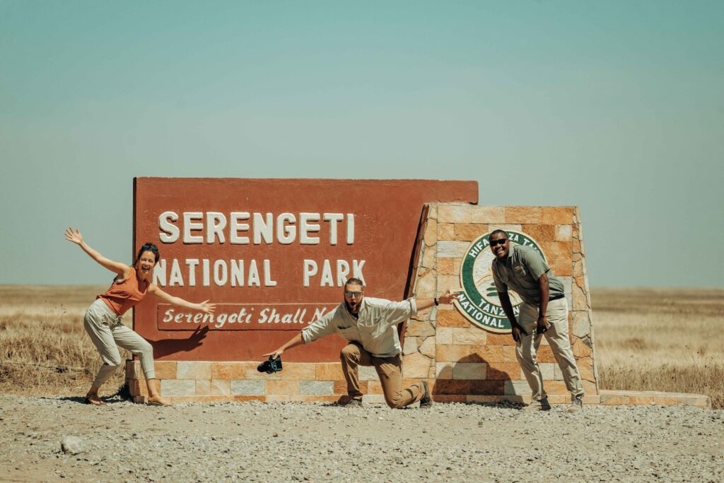 Three people pose enthusiastically in front of the Serengeti National Park entrance sign in Tanzania, with a dry, open landscape and clear blue sky—ready to explore the pristine trails that change with the seasons.