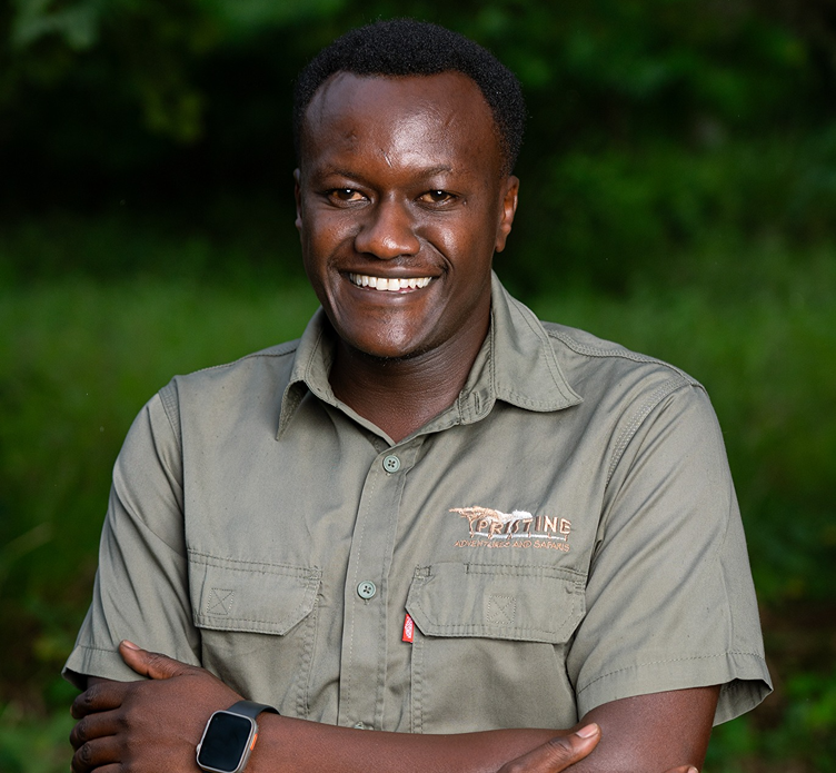 Meet the Team: A man in a green button-up shirt stands outdoors with arms crossed, smiling at the camera. He wears a smartwatch, framed by lush green foliage in the background.