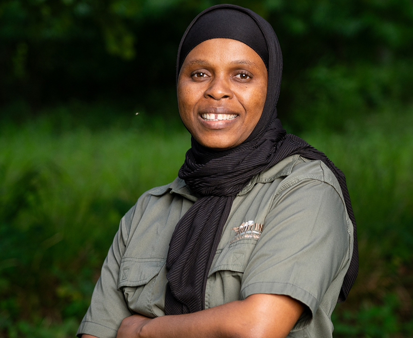 Meet The Team: A woman wearing a black headscarf and a grey collared shirt stands outdoors with her arms folded, smiling at the camera. Green foliage is blurred in the background.