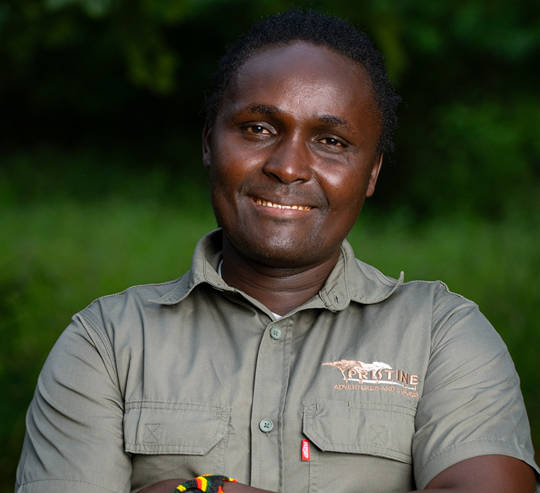 Meet The Team: A person stands outdoors, smiling with arms crossed, wearing a khaki shirt with a "Pristine Adventures and Safaris" logo. Lush green foliage creates an inviting backdrop.