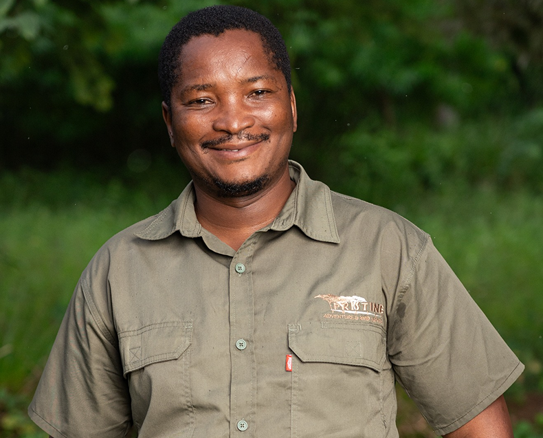 Meet The Team: Man standing outdoors, smiling in a khaki short-sleeve shirt with "PRISTINE" and an animal logo on the chest, green foliage in the background.