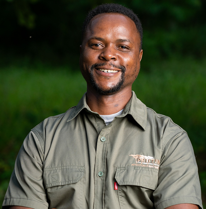 Meet The Team: A man in a green button-up shirt sits outdoors, smiling at the camera with a background of greenery.