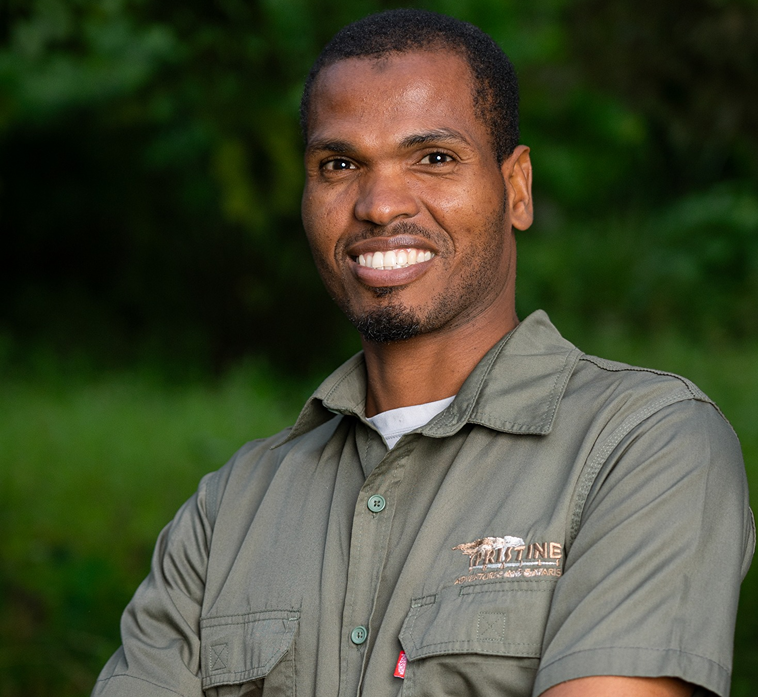 Meet the Team: A man in a green collared shirt with an embroidered logo stands outdoors, smiling confidently with arms crossed, surrounded by blurred greenery in the background.
