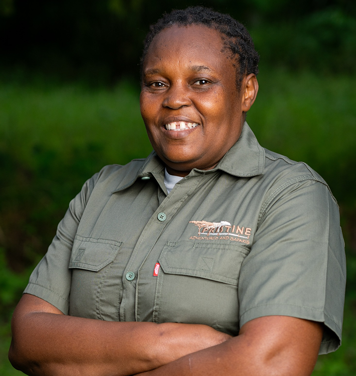 Meet the Team: A person in a green short-sleeve uniform shirt stands outdoors with arms crossed, smiling, surrounded by lush greenery in the background.