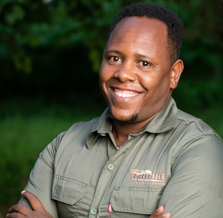 Meet The Team: A person in a khaki shirt with the "Pristine Trails and Safaris" logo stands outdoors, smiling with arms crossed against a green, blurred background.