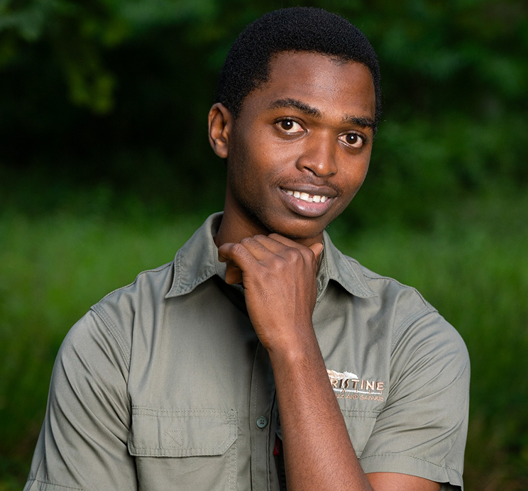 Meet the Team: A young man in a khaki shirt stands outdoors, facing the camera with one hand resting under his chin and greenery in the background.