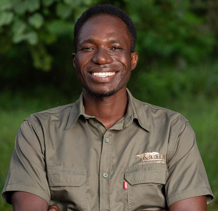 Meet the Team: A man in a green button-up shirt stands outdoors with his arms crossed, smiling at the camera. Green foliage is visible in the background.