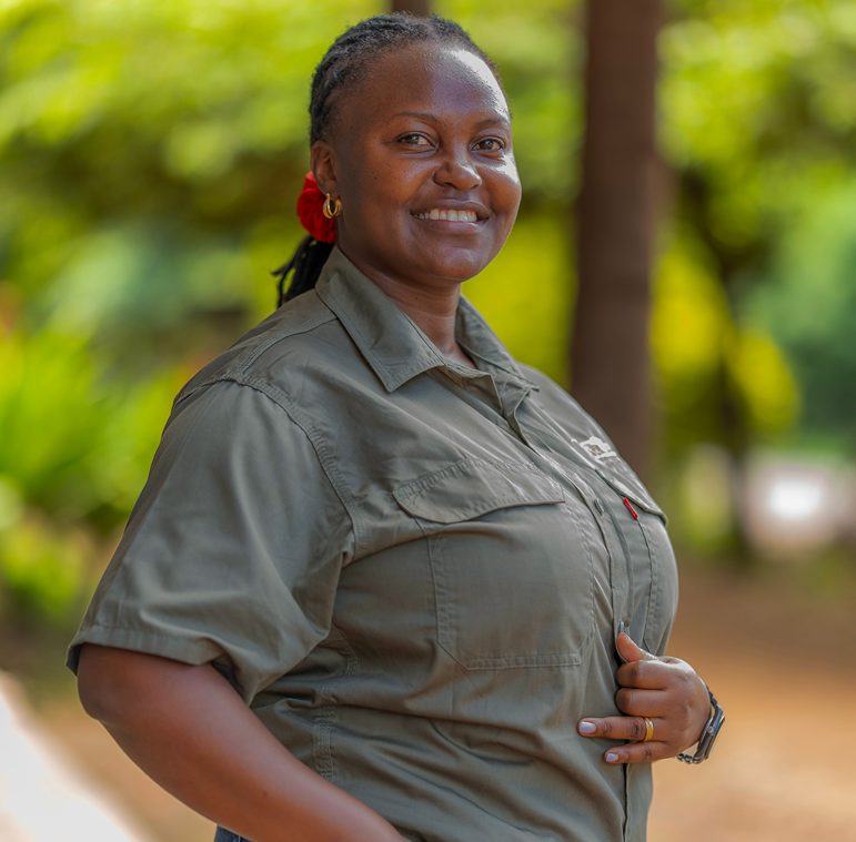 Meet The Team: A person in a green button-up shirt stands outdoors, smiling at the camera with greenery and trees in the blurred background.
