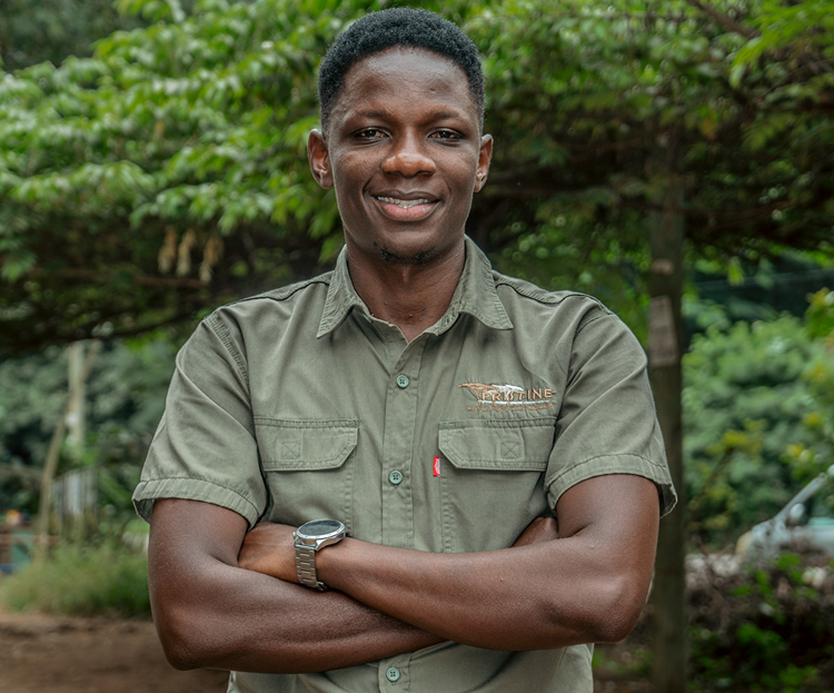 Meet The Team: A person stands outdoors with arms crossed, wearing a green short-sleeved button-up shirt and a wristwatch, with trees and greenery in the background.
