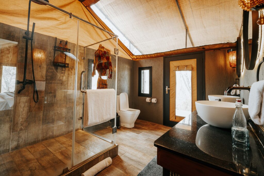 A modern bathroom inside a Nimali Tarangire luxury safari tent, featuring a glass-enclosed shower, a toilet, and double basins.