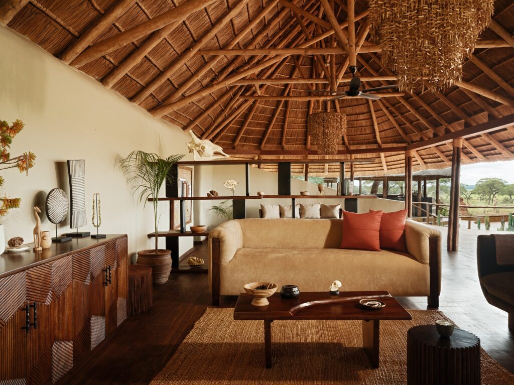 A living room at Sanctuary Swala features a tan sofa with two red scatter cushions facing a wooden coffee table under a circular thatched roof.