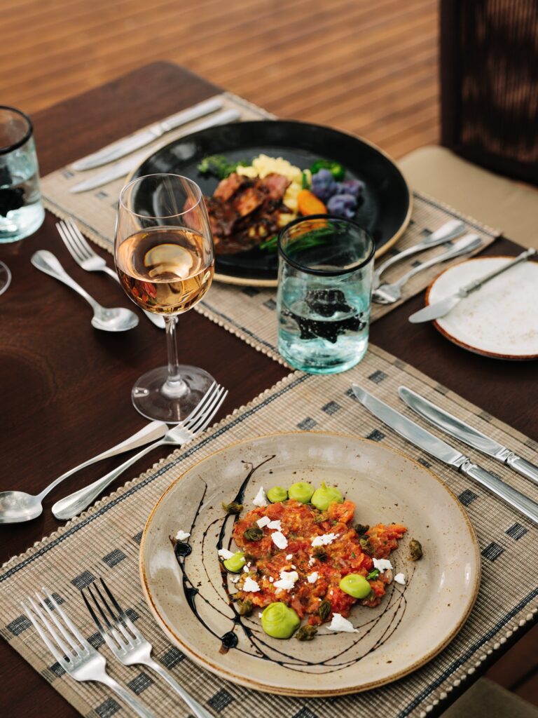 A tomato-based starter topped with cheese, capers, and herb purée, paired with rosé wine at a Sanctuary Swala luxury safari, with a main course on a wooden table.