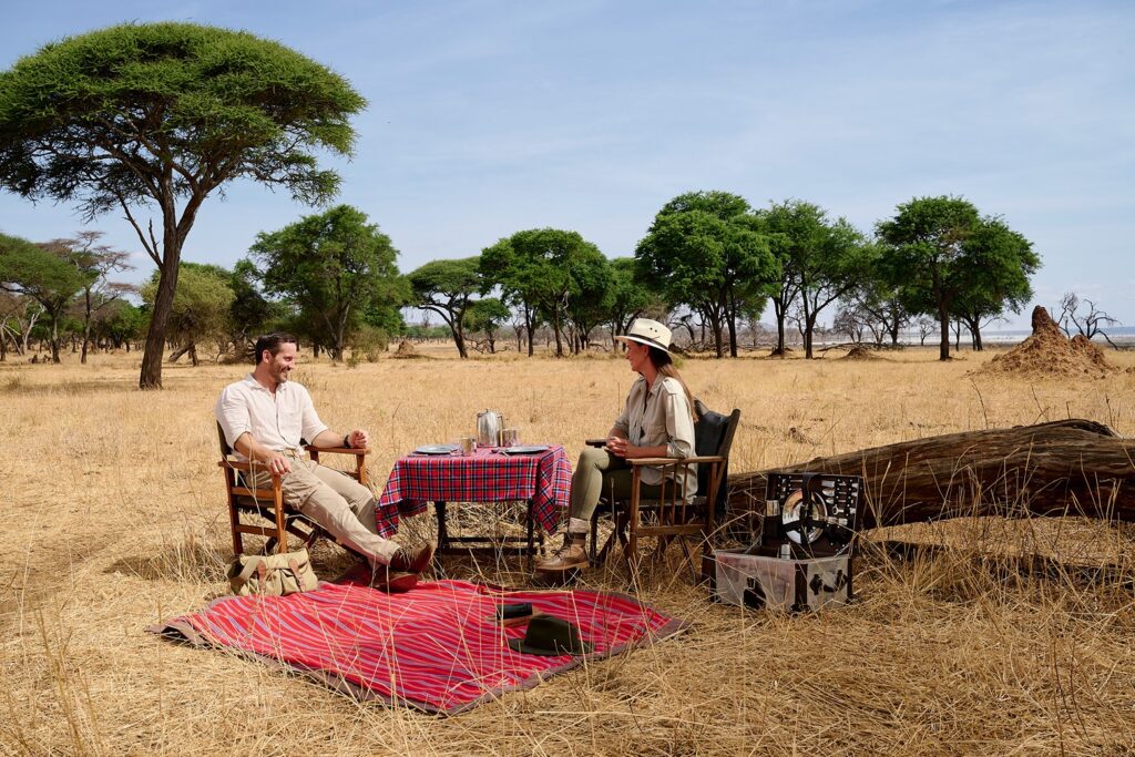 A man and a woman sit in folding chairs at a small table set for tea during a Tanzania safari on the grassy savannah.